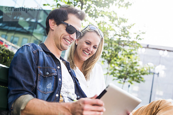 Glückliches junges Paar sitzt auf der Bank und schaut auf das digitale Tablett
