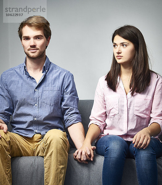 Seriöses junges Paar  das auf der Couch sitzt und Händchen hält.
