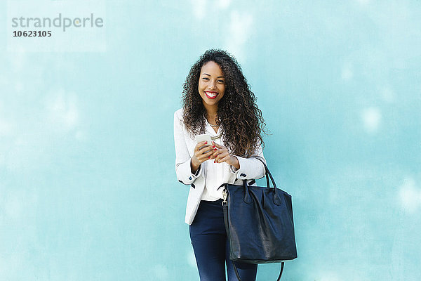 Porträt einer lächelnden jungen Geschäftsfrau mit Smartphone und Ledertasche vor einer blauen Wand