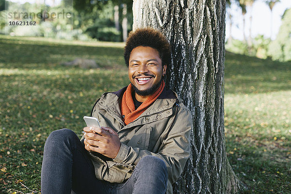 Porträt eines lächelnden jungen Mannes mit Smartphone  das sich an den Baumstamm lehnt