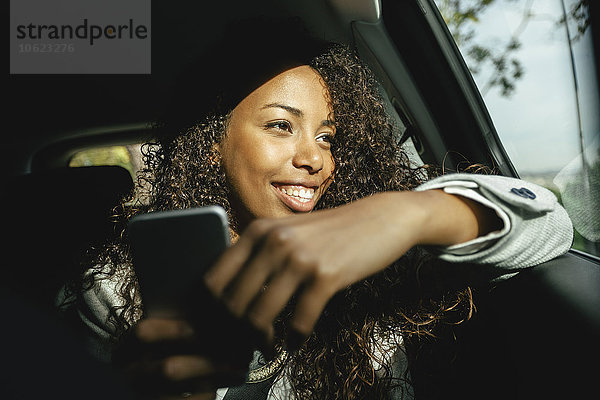 Lächelnde junge Frau sitzt in einem Auto mit Smartphone  das durchs Fenster schaut.