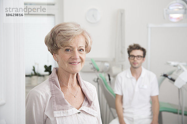 Porträt einer lächelnden Seniorin in der Chirurgie mit Zahnarzt im Hintergrund