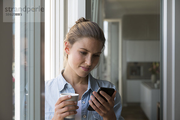 Junge Frau am Fenster stehend mit Smartphone und Kaffeetasse
