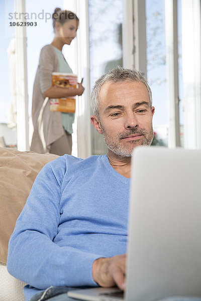 Mann auf Couch sitzend mit Laptop  Frau im Hintergrund