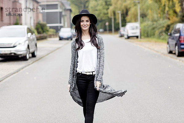 Porträt einer lächelnden jungen Frau mit schwarzem Hut  die auf einer Straße läuft.