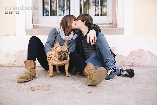 Junges Paar mit französischer Bulldogge auf dem Boden sitzend und küssend