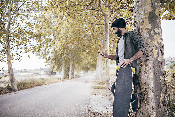 Spanien  Tarragona  junger Mann mit Longboard an Baumstamm lehnend auf sein Smartphone schauend