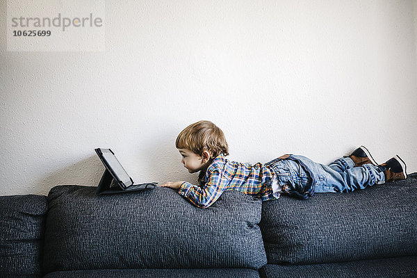 Kleiner Junge liegt auf der Rückenlehne einer Couch und schaut auf ein digitales Tablett.