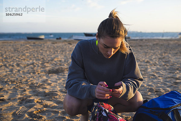 Spanien  Puerto Real  junge Frau  die auf ihr Smartphone schaut  während sie am Strand hockt