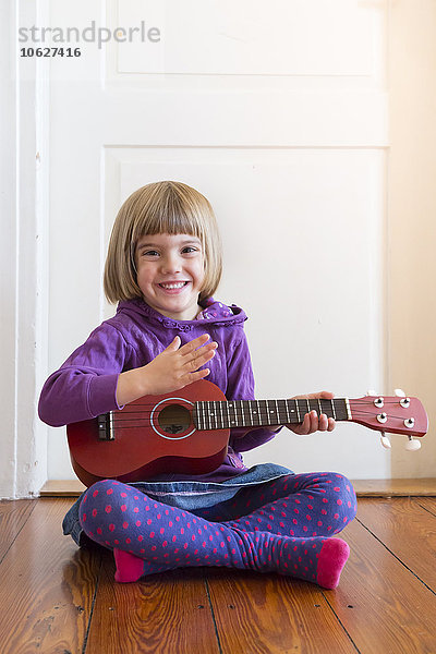 Porträt eines kleinen Mädchens  das auf einem Holzboden sitzt und Ukulelele spielt.