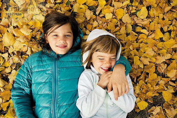 Zwei Mädchen liegen nebeneinander auf einem mit Herbstlaub bedeckten Boden.