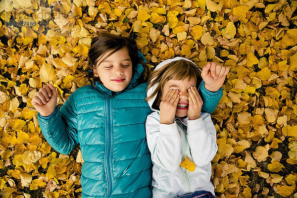 Zwei Mädchen liegen nebeneinander auf einem mit Herbstlaub bedeckten Boden.