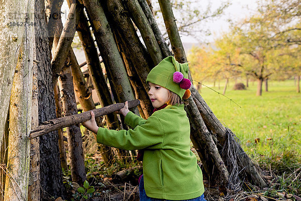 Kleines Mädchen beim Bau einer Hütte mit Baumstämmen auf einer Wiese im Herbst