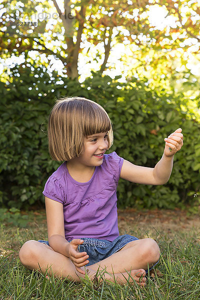 Lächelndes kleines Mädchen  das auf einer Wiese sitzt und etwas in der Hand hält.