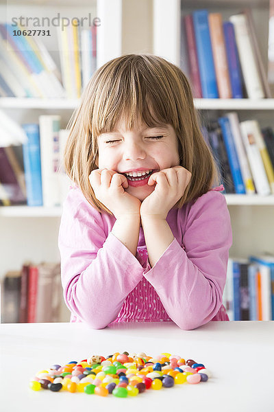 Porträt eines lächelnden kleinen Mädchens mit geschlossenen Augen und auf einem Tisch liegenden Geleebohnen.