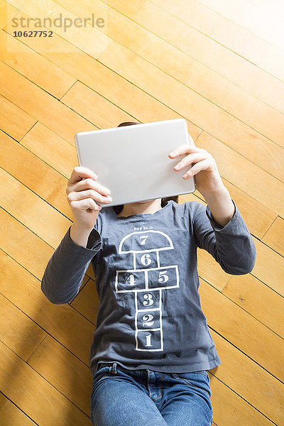 Mädchen liegt auf Holzboden und bedeckt ihr Gesicht mit einem digitalen Tablett.