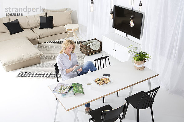 Lächelnde Frau sitzt am Tisch in ihrem Wohnzimmer mit Hilfe eines digitalen Tabletts.