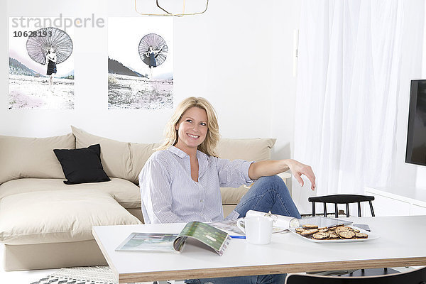 Porträt einer lächelnden Frau  die sich am Tisch in ihrem Wohnzimmer entspannt.