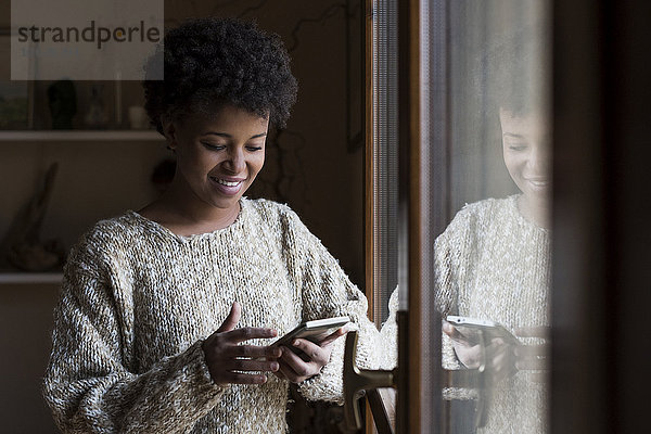 Lächelnde junge Frau am Fenster mit Blick auf das Smartphone