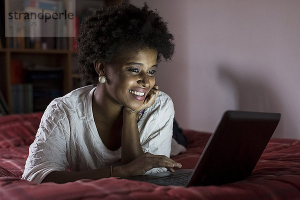 Porträt einer lächelnden jungen Frau auf dem Bett liegend mit Laptop