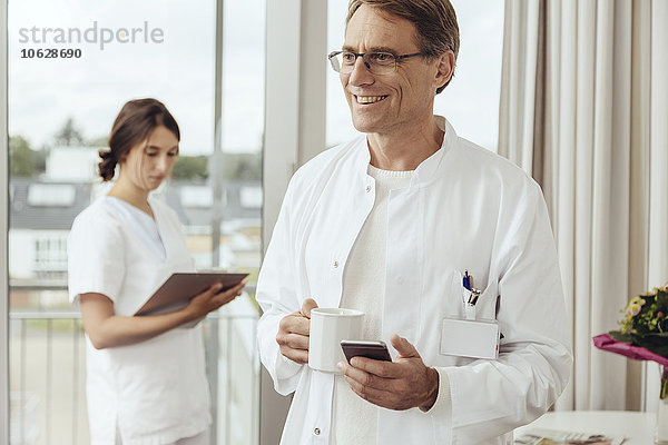 Arzt mit Kaffeetasse und Smartphone  Krankenschwester arbeitet im Hintergrund