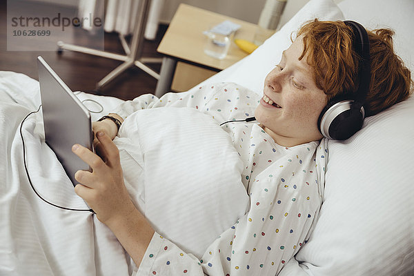 Kranker Junge  der im Krankenhaus liegt  mit digitalem Tablett und Kopfhörern.