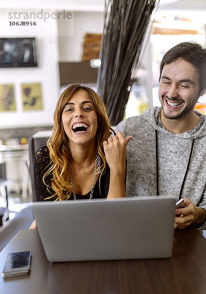 Porträt eines lachenden jungen Paares mit Laptop in einer Kneipe