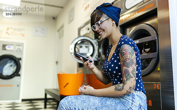 Portrait der tätowierten jungen Frau mit Smartphone im Waschsalon