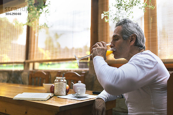 Mann sitzt am Frühstückstisch und trinkt ein Glas Saft.