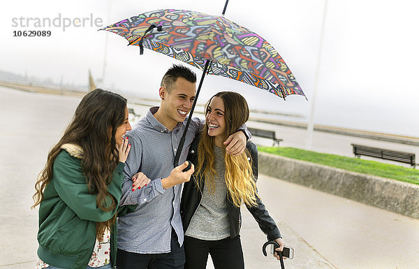 Drei glückliche Freunde  die mit einem Regenschirm die Straße hinuntergehen.