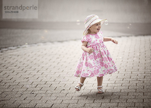 Fröhliches blondes kleines Mädchen mit Hut und Sommerkleid mit Blumenmuster  das auf dem Bürgersteig tanzt.