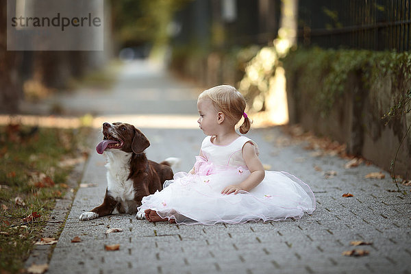 Blondes kleines Mädchen im Tüllkleid neben ihrem Hund auf dem Bürgersteig sitzend