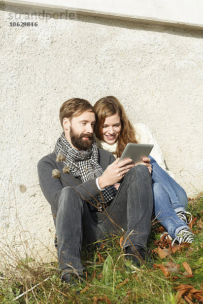 Lächelndes Paar sitzt auf der Wiese und schaut auf ein digitales Tablett.