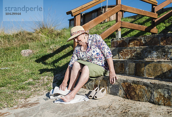 Seniorin sitzt auf einer Treppe und wischt sich den Sand von den Füßen.