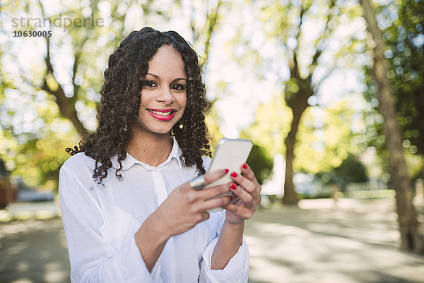 Porträt einer lächelnden jungen Frau mit braunen Ringeln und roten Lippen mit Smartphone