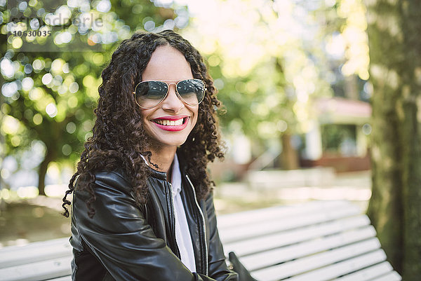Porträt einer lächelnden Frau mit Sonnenbrille und Lederjacke  die auf einer Parkbank sitzt.