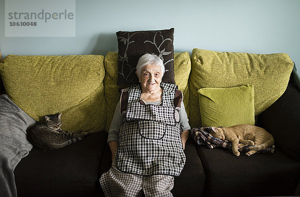 Porträt einer älteren Frau  die zu Hause mit ihren beiden schlafenden Katzen auf der Couch sitzt.