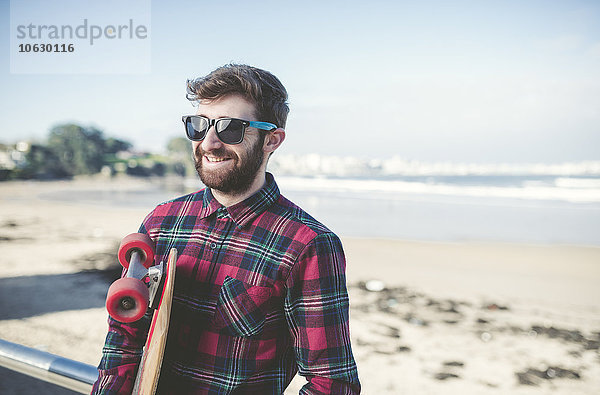 Spanien  La Coruna  Porträt eines lächelnden Hipsters mit Sonnenbrille und Skateboard am Strand