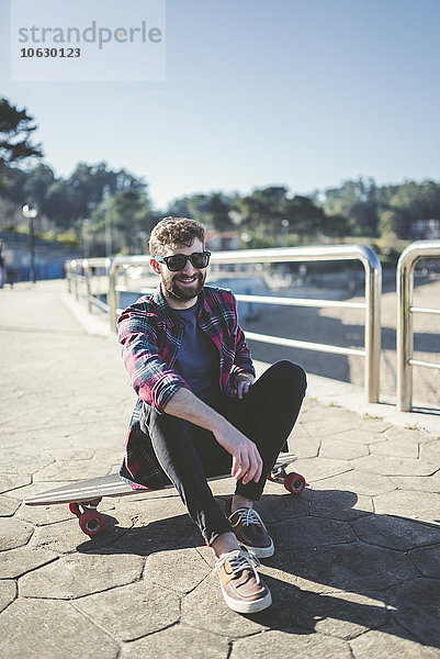 Spanien  La Coruna  Porträt eines lächelnden Hipsters mit Sonnenbrille auf dem Longboard