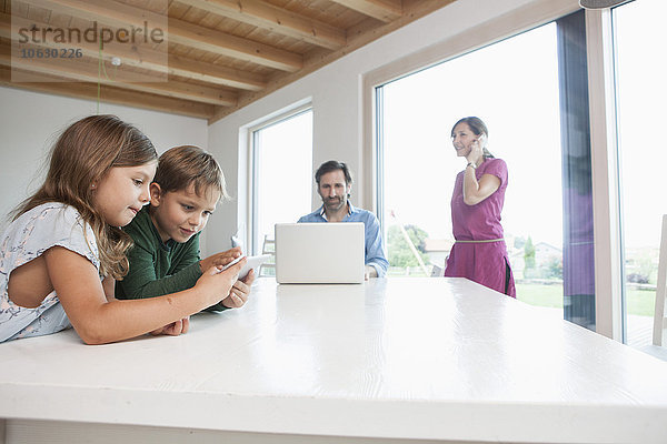 Geschwister mit digitalem Tablett  Eltern mit Laptop im Hintergrund