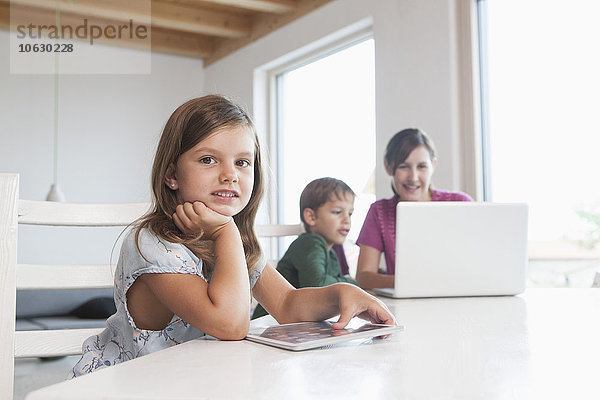 Kleines Mädchen mit digitalem Tablett  Mutter und Bruder mit Laptop im Hintergrund