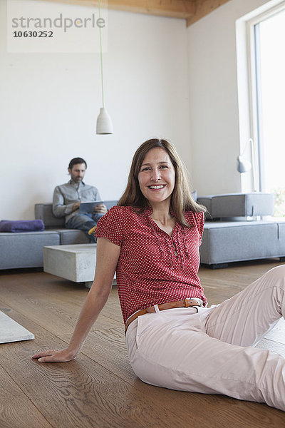Reife Frau lächelnd auf dem Boden im Wohnzimmer sitzend  Mann mit digitalem Tablett im Hintergrund