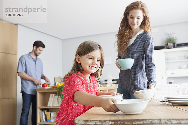 Lächelndes kleines Mädchen deckt den Tisch in der Küche mit ihren Eltern im Hintergrund.