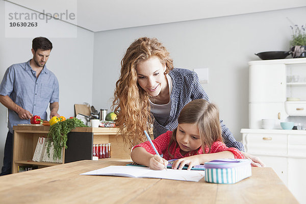 Kleines Mädchen bei den Hausaufgaben am Küchentisch mit ihren Eltern im Hintergrund