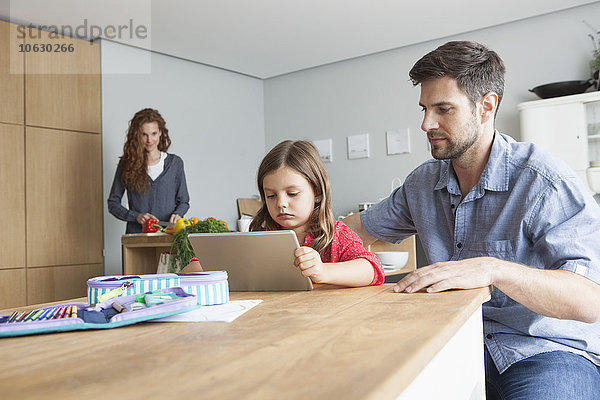 Kleines Mädchen und ihr Vater sitzen am Küchentisch und schauen auf ein digitales Tablett.