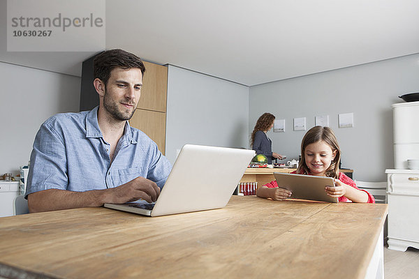 Mann und seine kleine Tochter sitzen am Küchentisch mit Laptop und digitalem Tablett