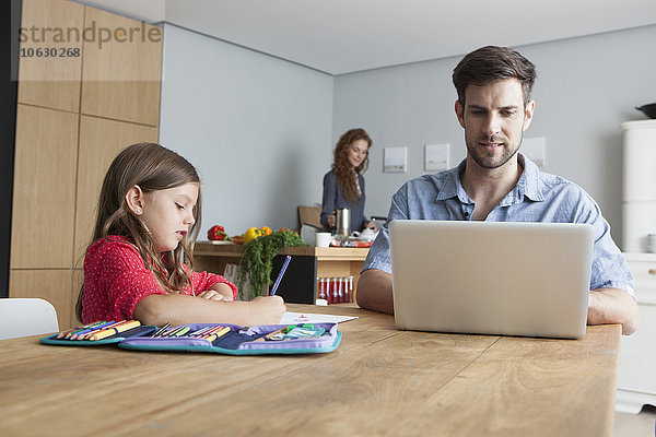 Mann sitzt am Küchentisch mit Laptop  während seine kleine Tochter malt.