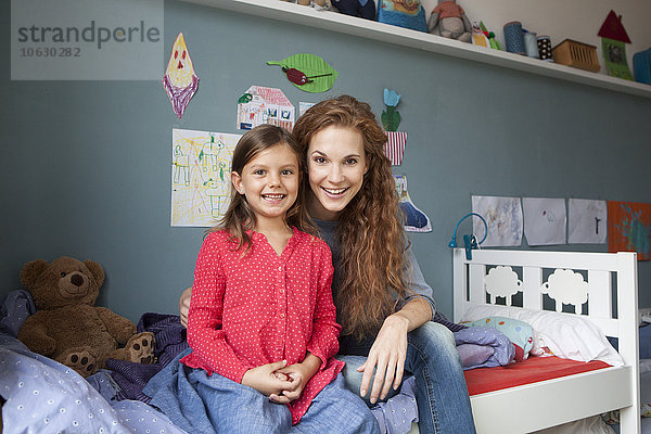 Porträt der Mutter und der kleinen Tochter  die nebeneinander auf dem Bett im Kinderzimmer sitzen.