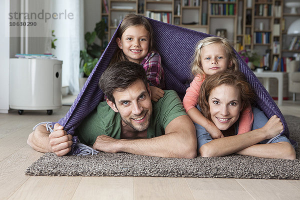 Porträt eines Paares mit ihren kleinen Töchtern  die zusammen auf dem Boden des Wohnzimmers liegen und von einer Decke bedeckt sind.