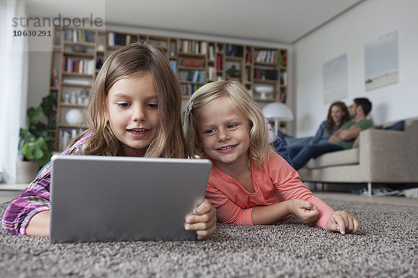 Porträt von zwei kleinen Schwestern auf dem Boden liegend im Wohnzimmer mit digitalem Tablett